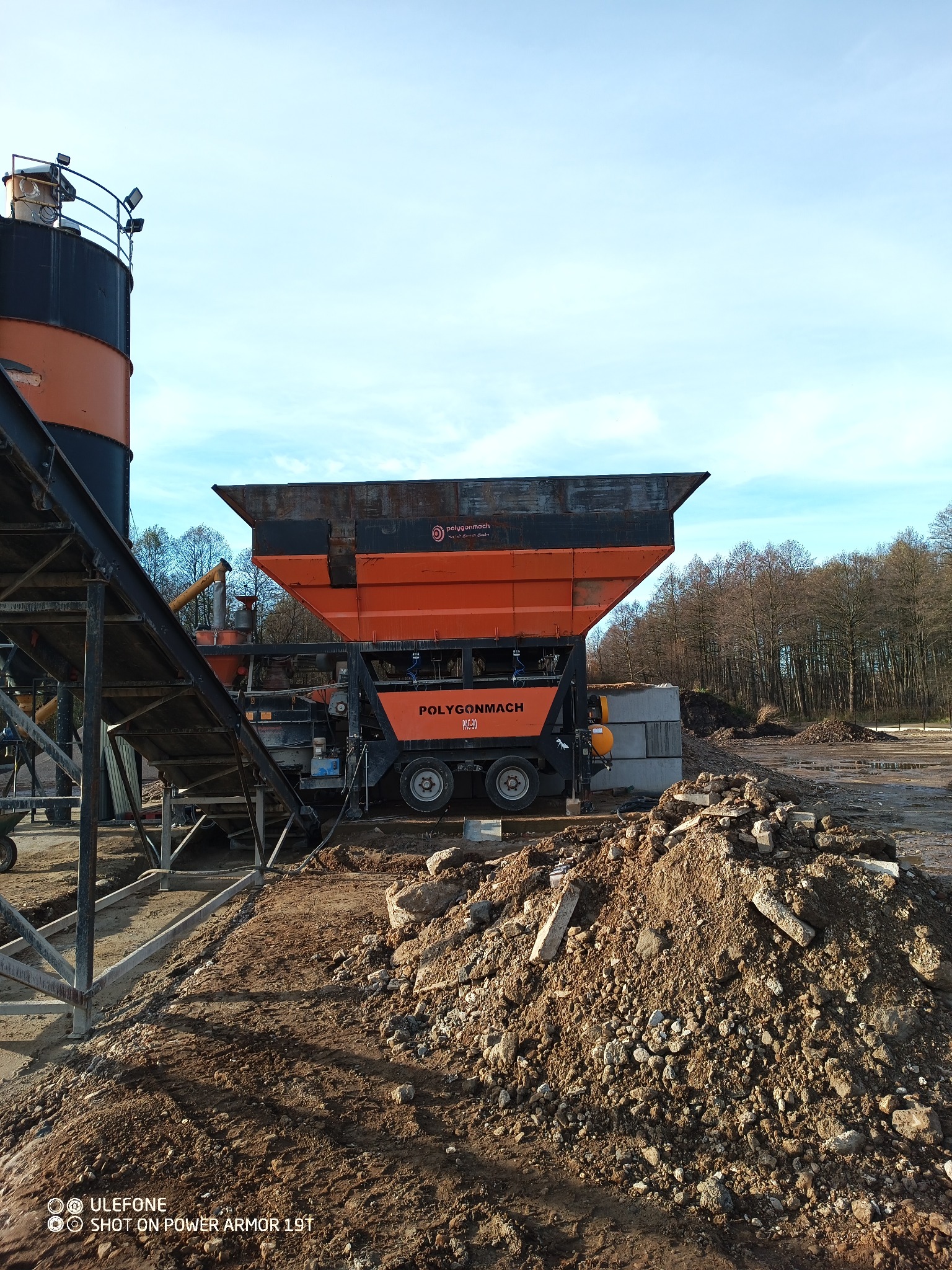 Pomarańczowo-czarna maszyna do obróbki kruszyw marki Polygonmach na placu budowy, z widoczną konstrukcją stalową i stertą ziemi. Błękitne niebo w tle.