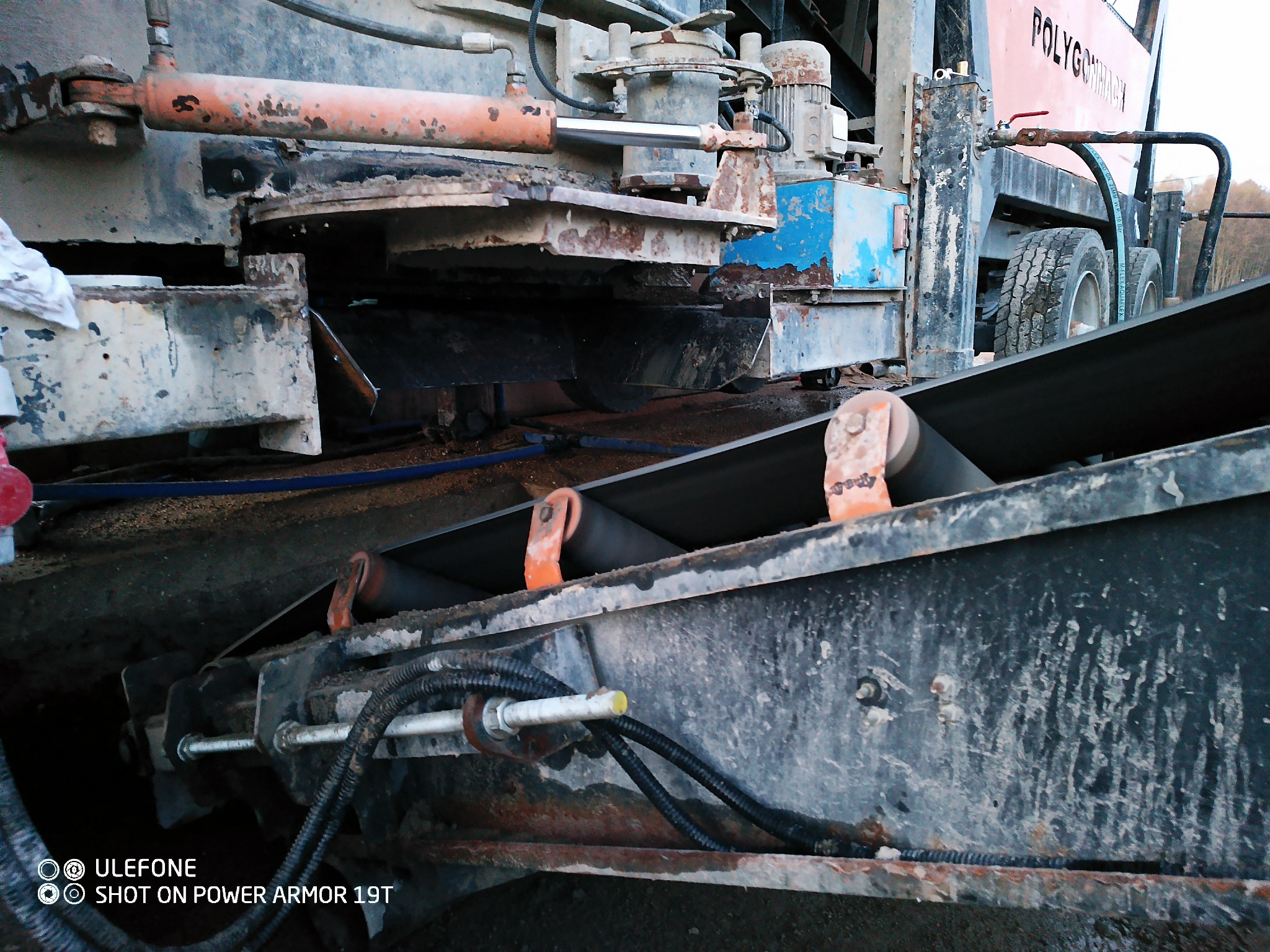 Fragment maszyny budowlanej z widocznym przenośnikiem taśmowym i elementami hydraulicznymi, oświetlony naturalnym światłem, z widocznymi śladami użytkowania.