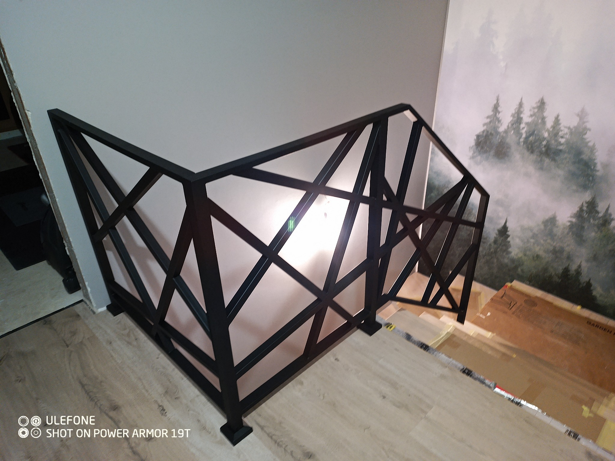 Czarna, metalowa balustrada schodowa o geometrycznym wzorze, zamontowana wewnątrz budynku. W tle ściana z tapetą w motywie leśnym i drewniane schody.