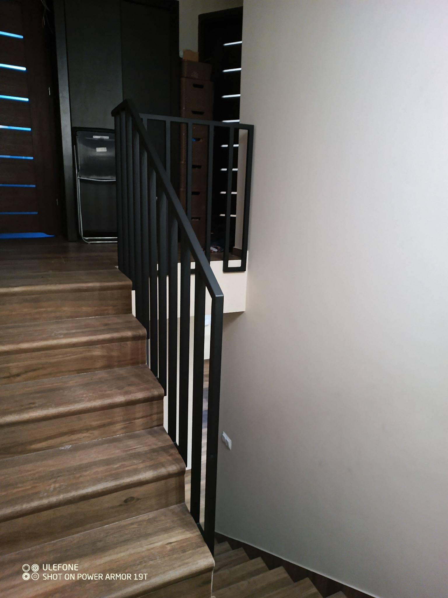 Drewniane schody z czarną, minimalistyczną balustradą z pionowych prętów. Widok z góry na dół, z fragmentem drzwi i szafy w tle. Nowoczesny design.