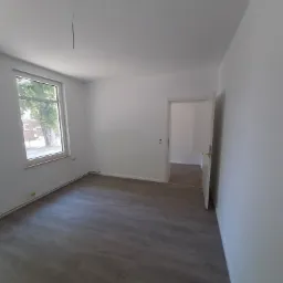 Pusty, odświeżony pokój z panelami podłogowymi w odcieniach szarości, białymi ścianami, grzejnikiem przypodłogowym i otwartymi drzwiami do sąsiedniego pomieszczenia, widok z okna na drzewo.