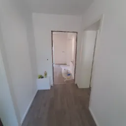 Wnętrze mieszkania w trakcie odświeżania: świeżo pomalowane ściany na biało, widoczne zabezpieczenia podłogi folią malarską, usunięte ościeżnice drzwiowe, butelka z płynem i narzędzia na p&oacute;łce.