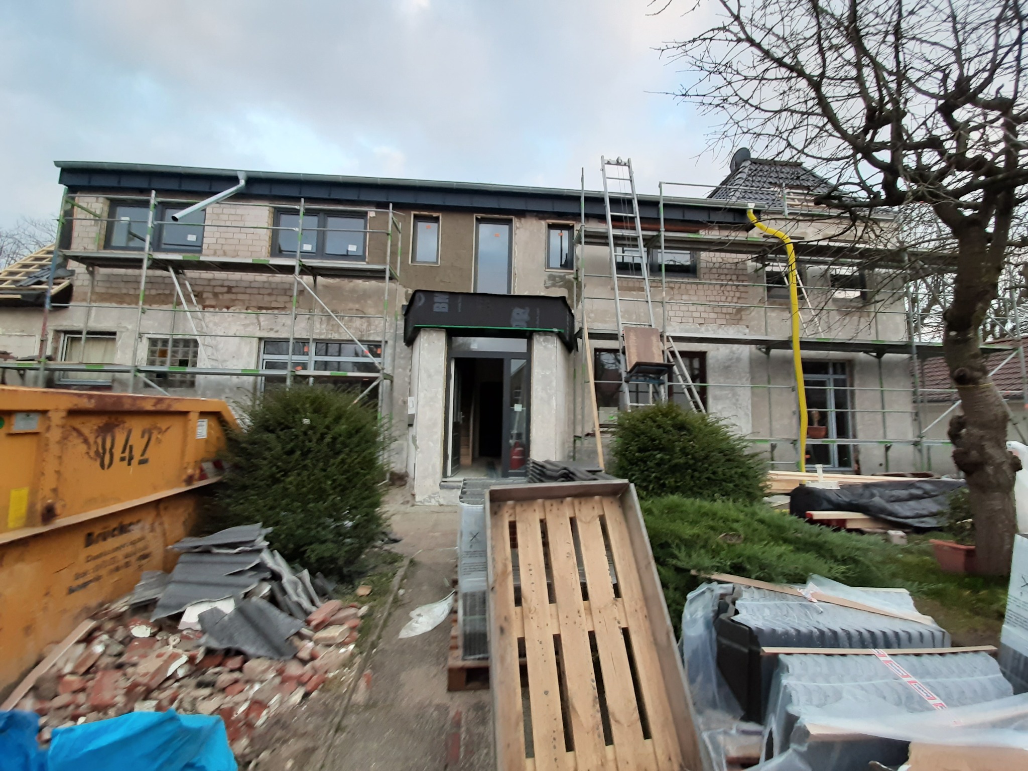 Dom w trakcie remontu elewacji, widoczne rusztowania, kontener na gruz i materiały budowlane, częściowo otynkowana ściana.