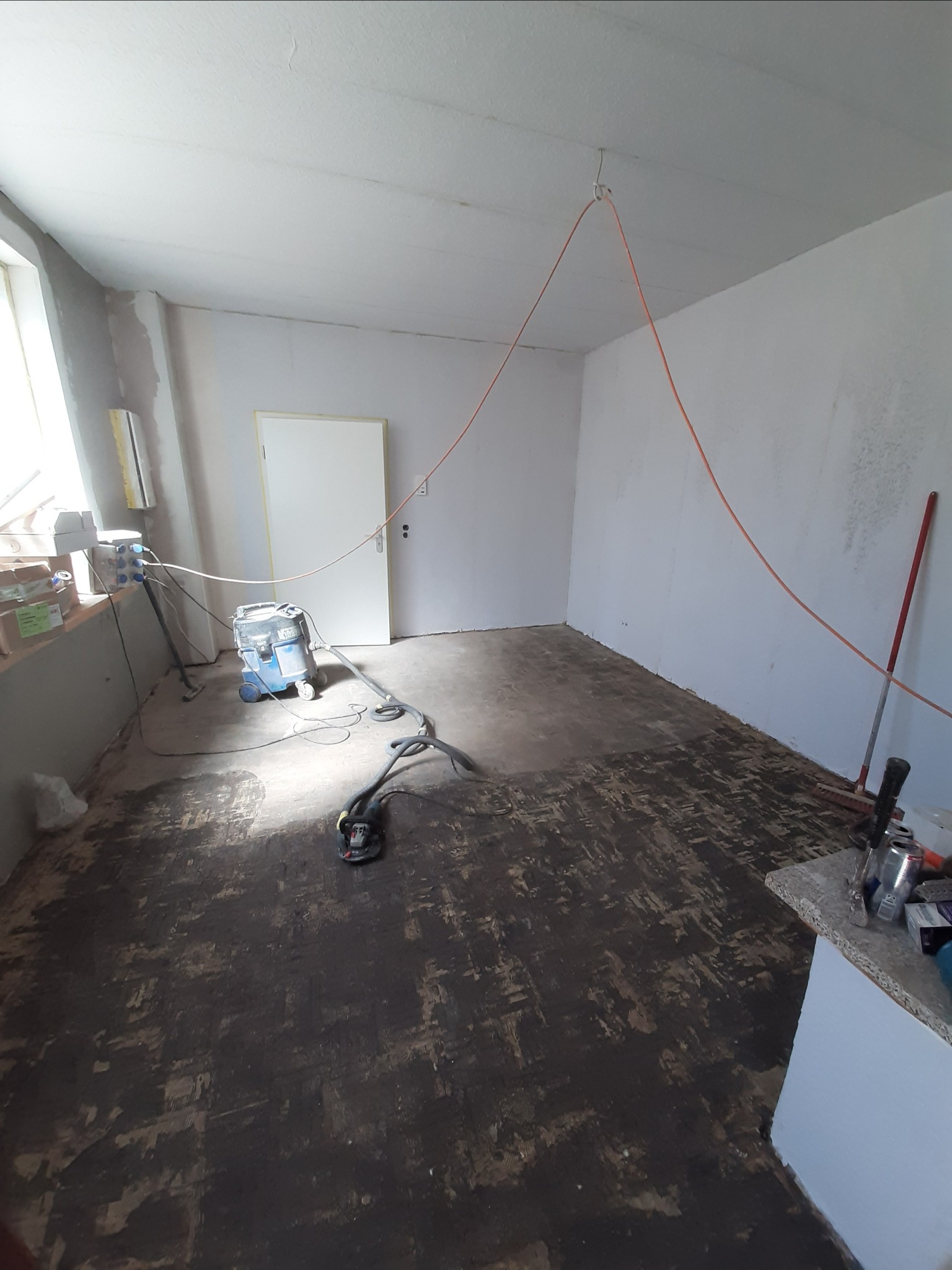 Przygotowanie podłoża w pomieszczeniu: szlifowanie posadzki za pomocą szlifierki podłączonej do odkurzacza przemysłowego, widoczne ślady po szlifowaniu, kabel podwieszony pod sufitem, białe ściany...