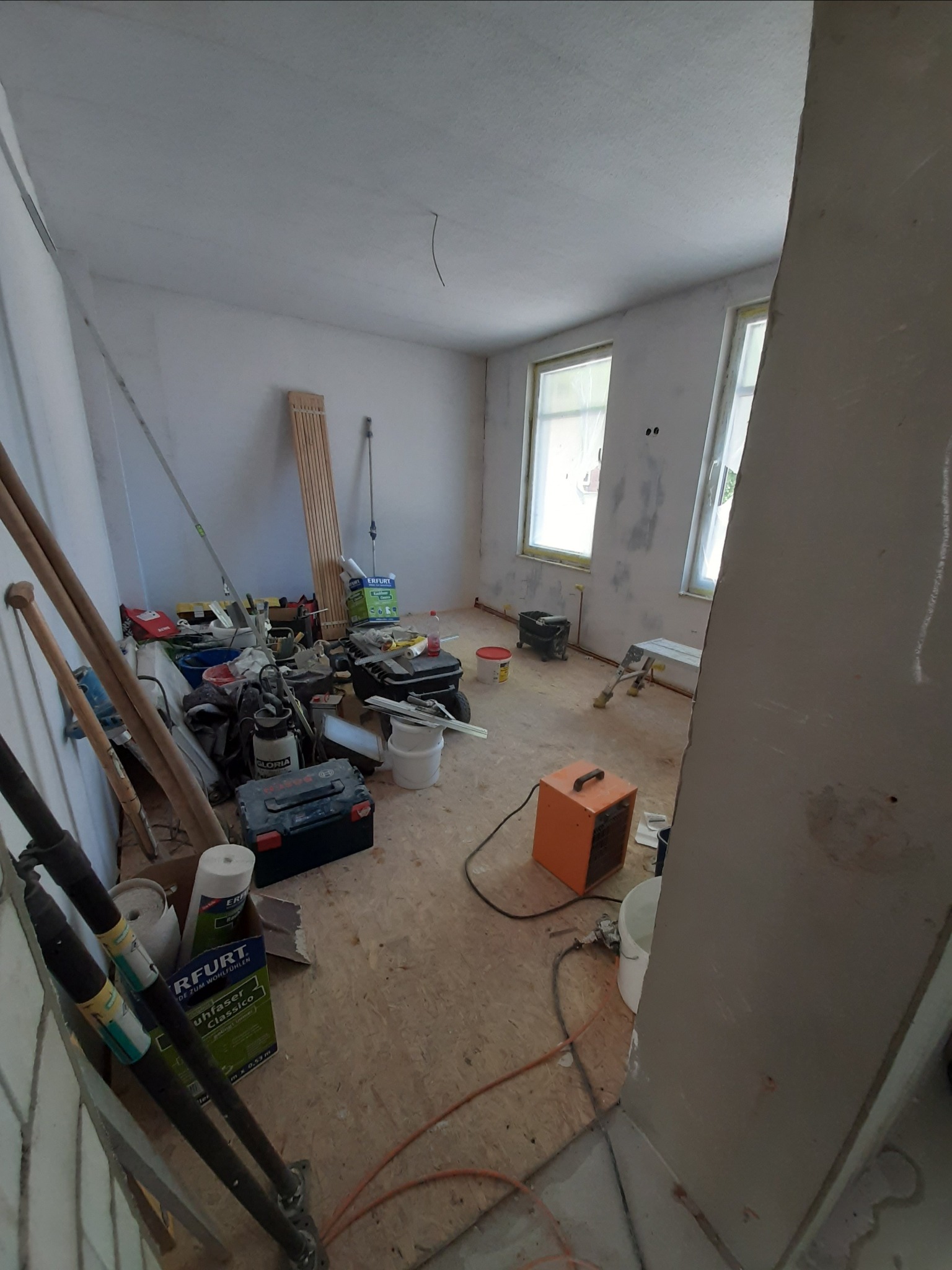 Pokój w trakcie prac wykończeniowych: ściany częściowo pomalowane, widoczne narzędzia, drabiny, wiadro z farbą, grzejnik elektryczny, oraz materiały budowlane na podłodze.