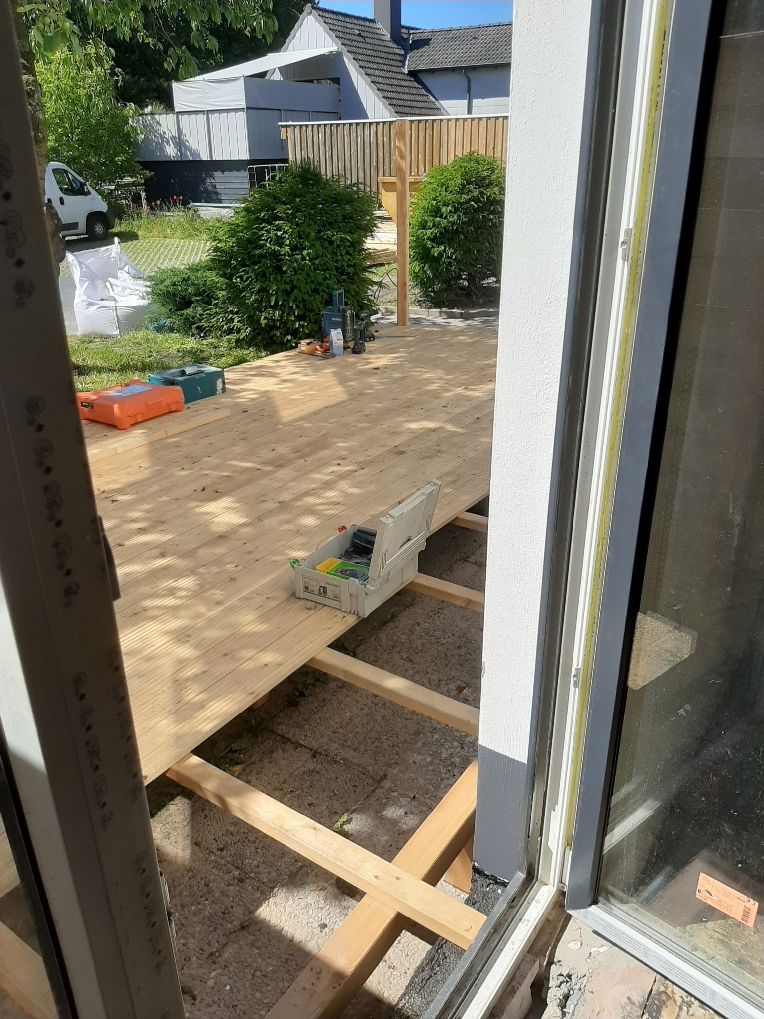 Montaż drewnianego tarasu: widok z wnętrza przez otwarte drzwi balkonowe na częściowo ułożone deski na legarach, skrzynka z narzędziami i akcesoriami na powierzchni tarasu, w tle zieleń i zabudowania.