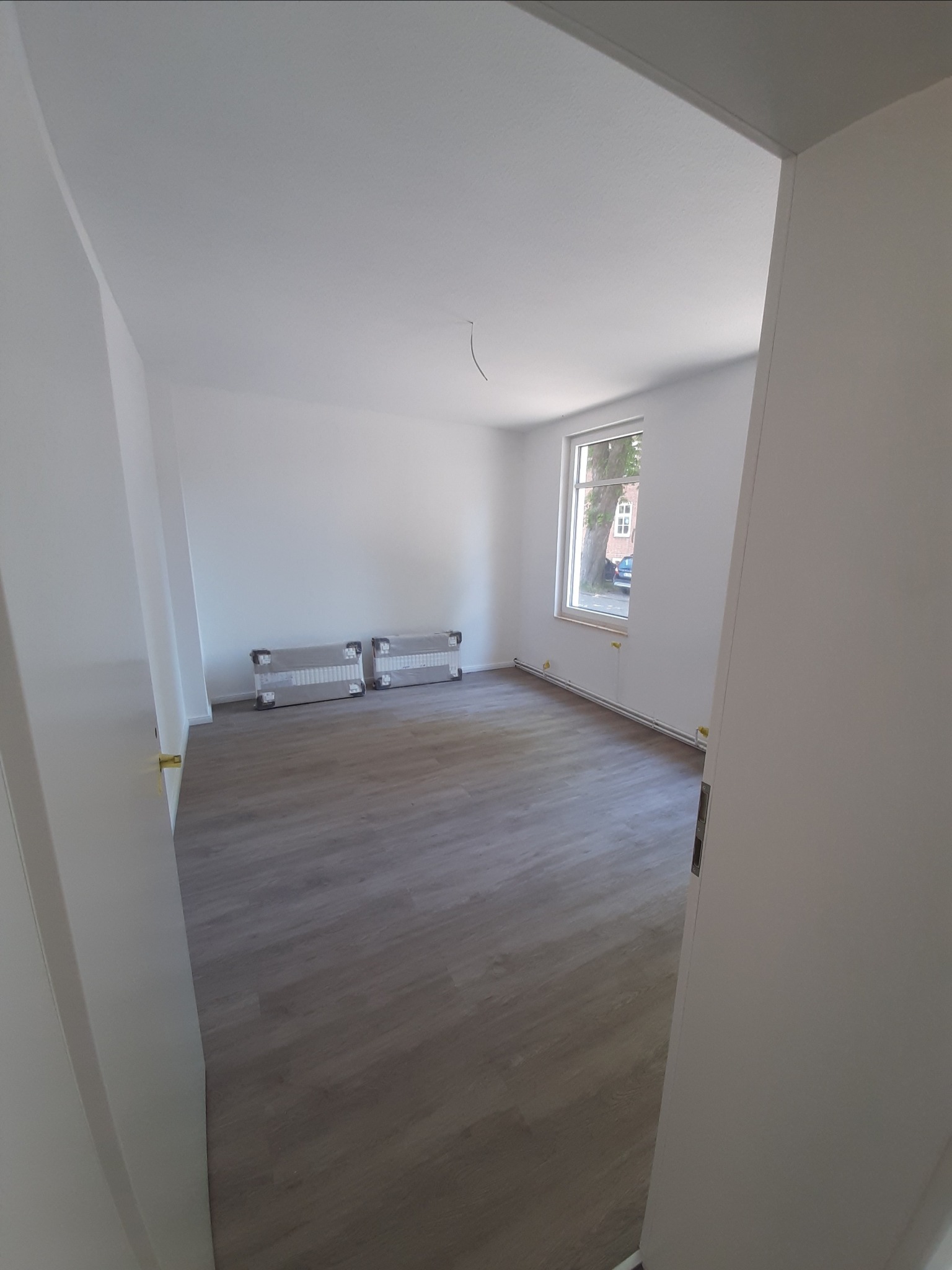Pusty pokój po remoncie z panelami podłogowymi, świeżo pomalowanymi ścianami na biało i dwoma grzejnikami owiniętymi folią ochronną. Widok zza otwartych drzwi.