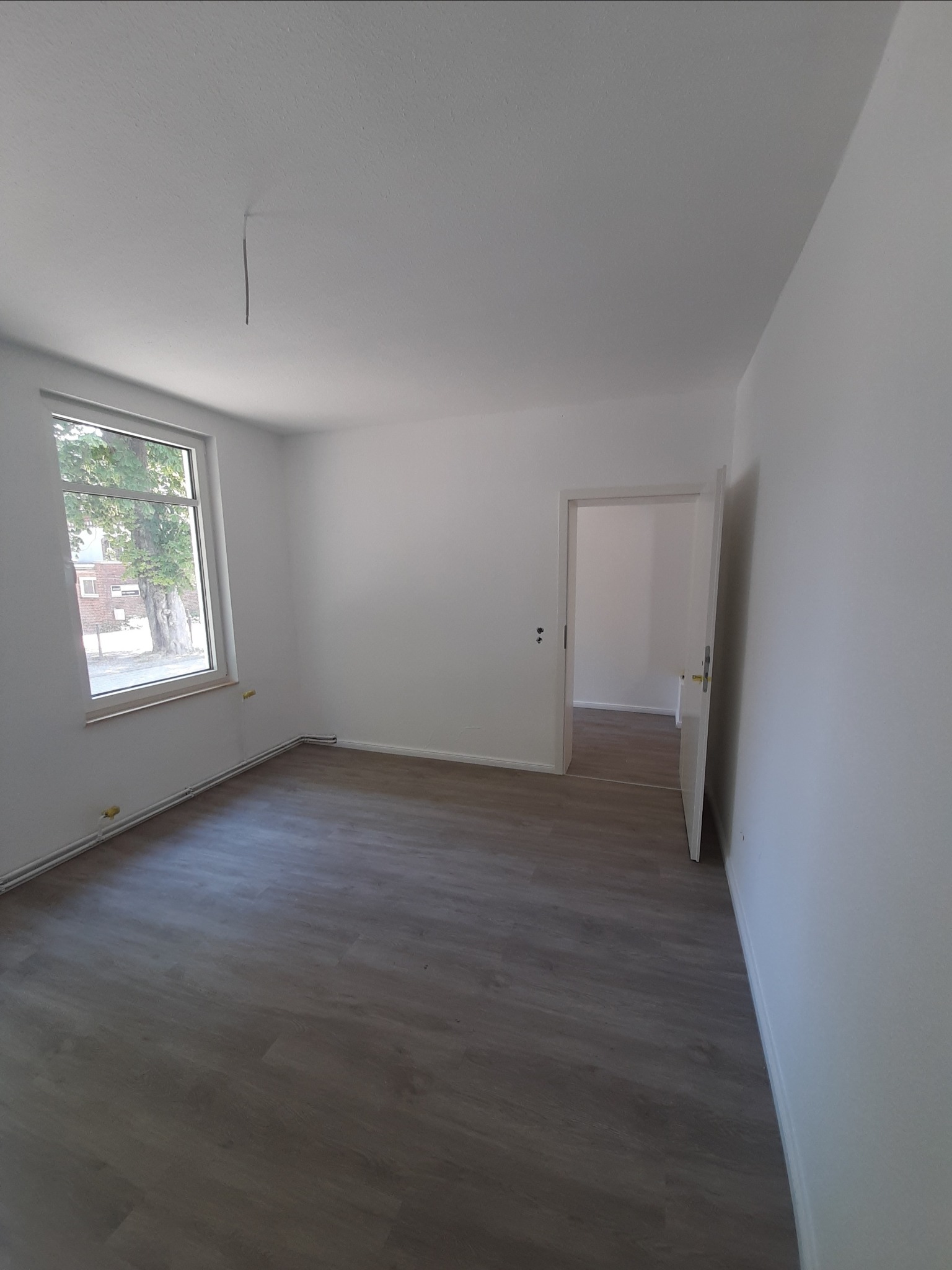 Pusty, odświeżony pokój z panelami podłogowymi w odcieniach szarości, białymi ścianami, grzejnikiem przypodłogowym i otwartymi drzwiami do sąsiedniego pomieszczenia, widok z okna na drzewo.