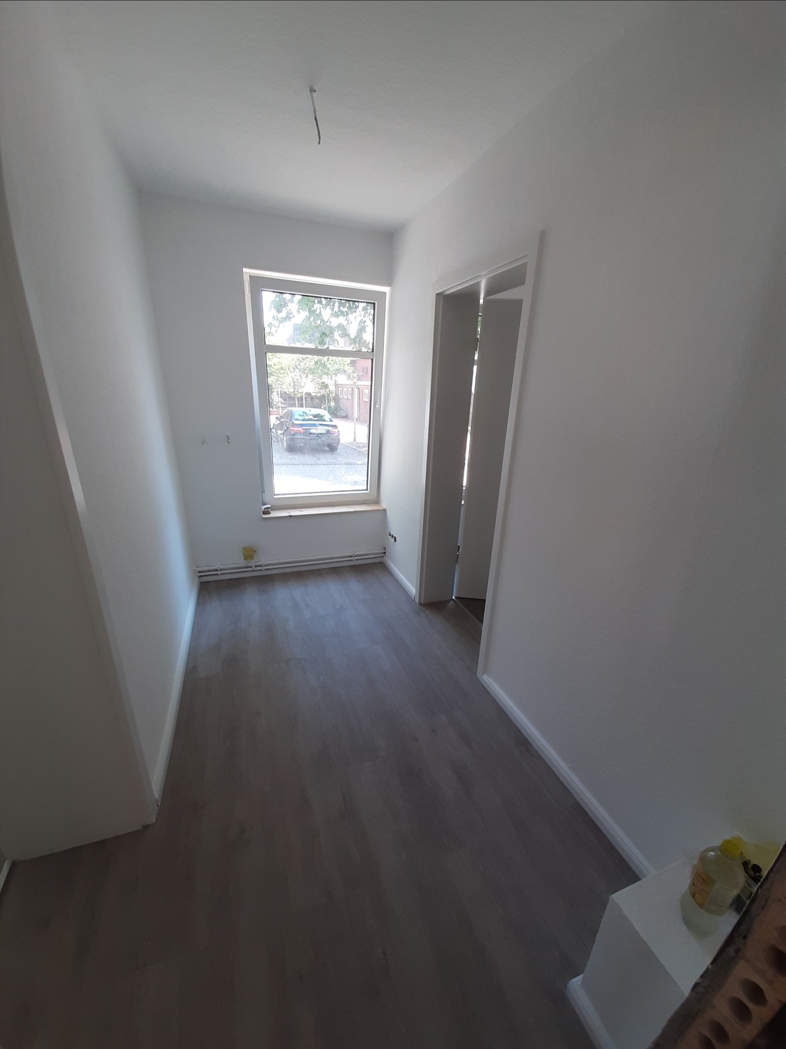 Wnętrze pokoju po remoncie w Choszcznie: świeżo pomalowane ściany na biało, szara podłoga, widok na ulicę przez okno.