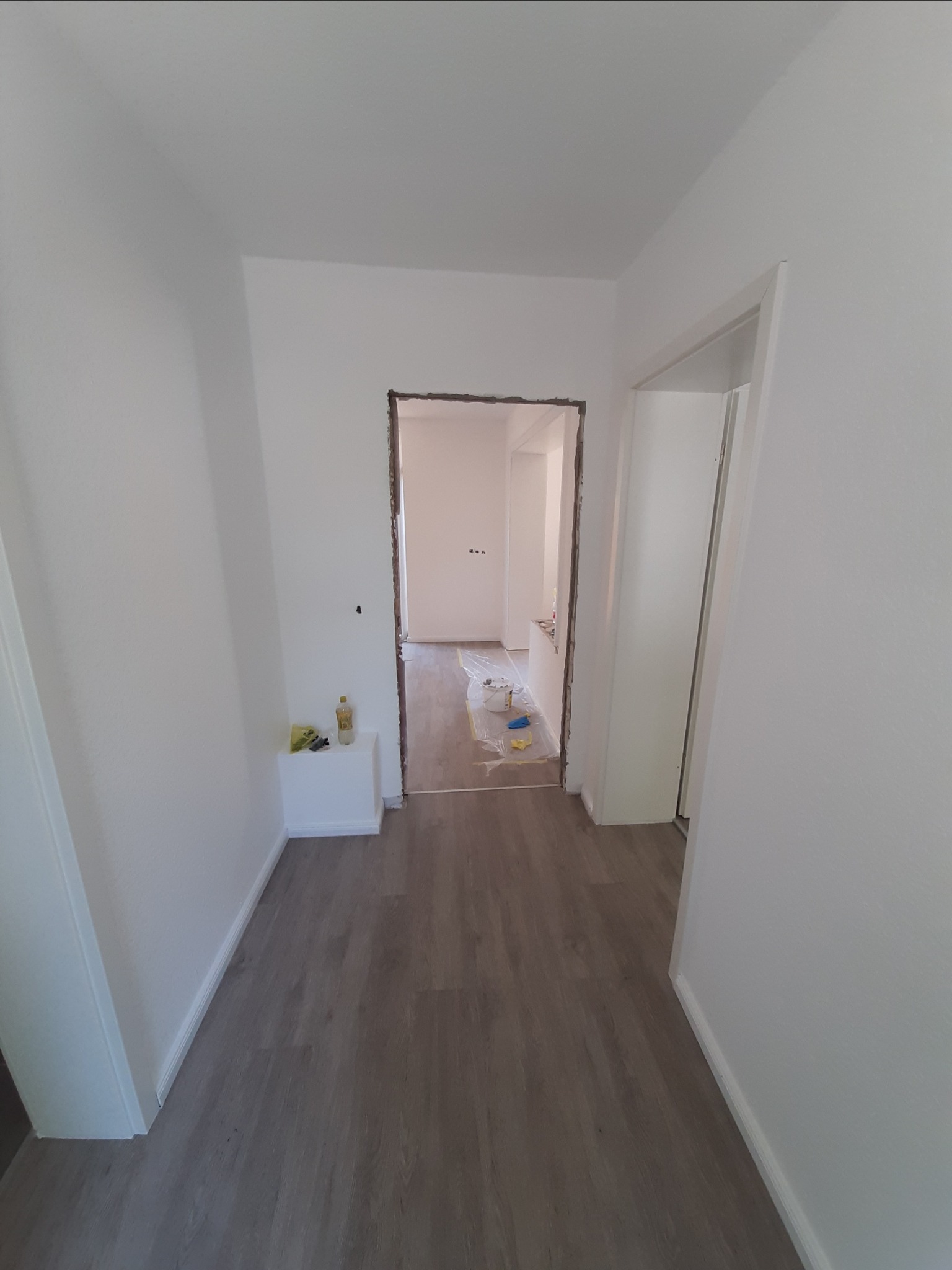 Wnętrze mieszkania w trakcie odświeżania: świeżo pomalowane ściany na biało, widoczne zabezpieczenia podłogi folią malarską, usunięte ościeżnice drzwiowe, butelka z płynem i narzędzia na półce.