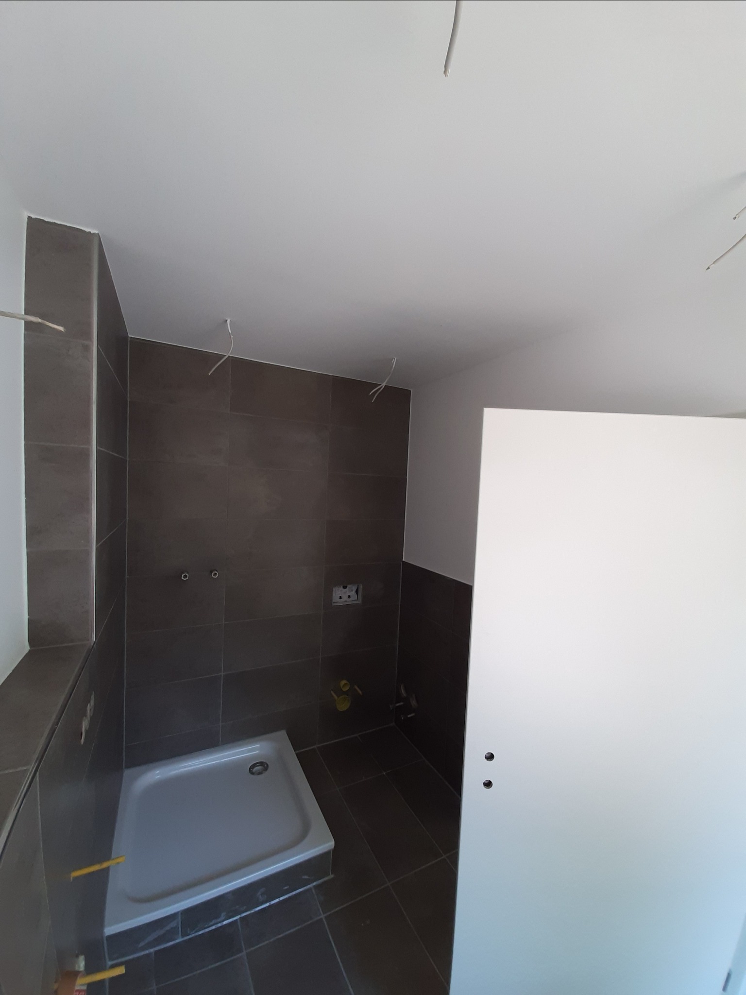 Niedokończona łazienka z ciemnymi płytkami, brodzikiem i częściowo zamontowanymi elementami instalacji, widoczne wystające kable.