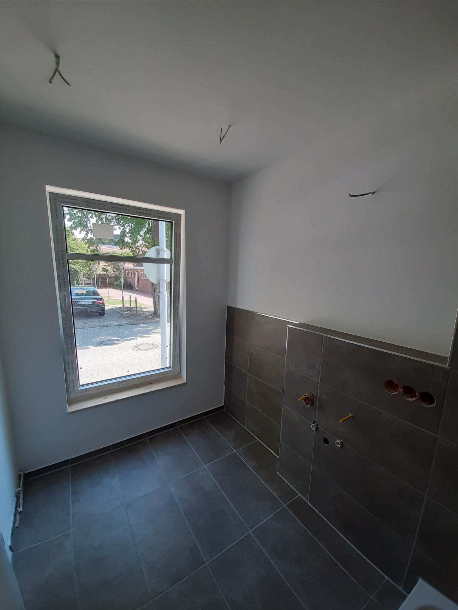 Wnętrze łazienki w trakcie remontu: szare płytki na podłodze i częściowo na ścianie, przygotowane punkty instalacyjne, widok na ulicę przez okno.