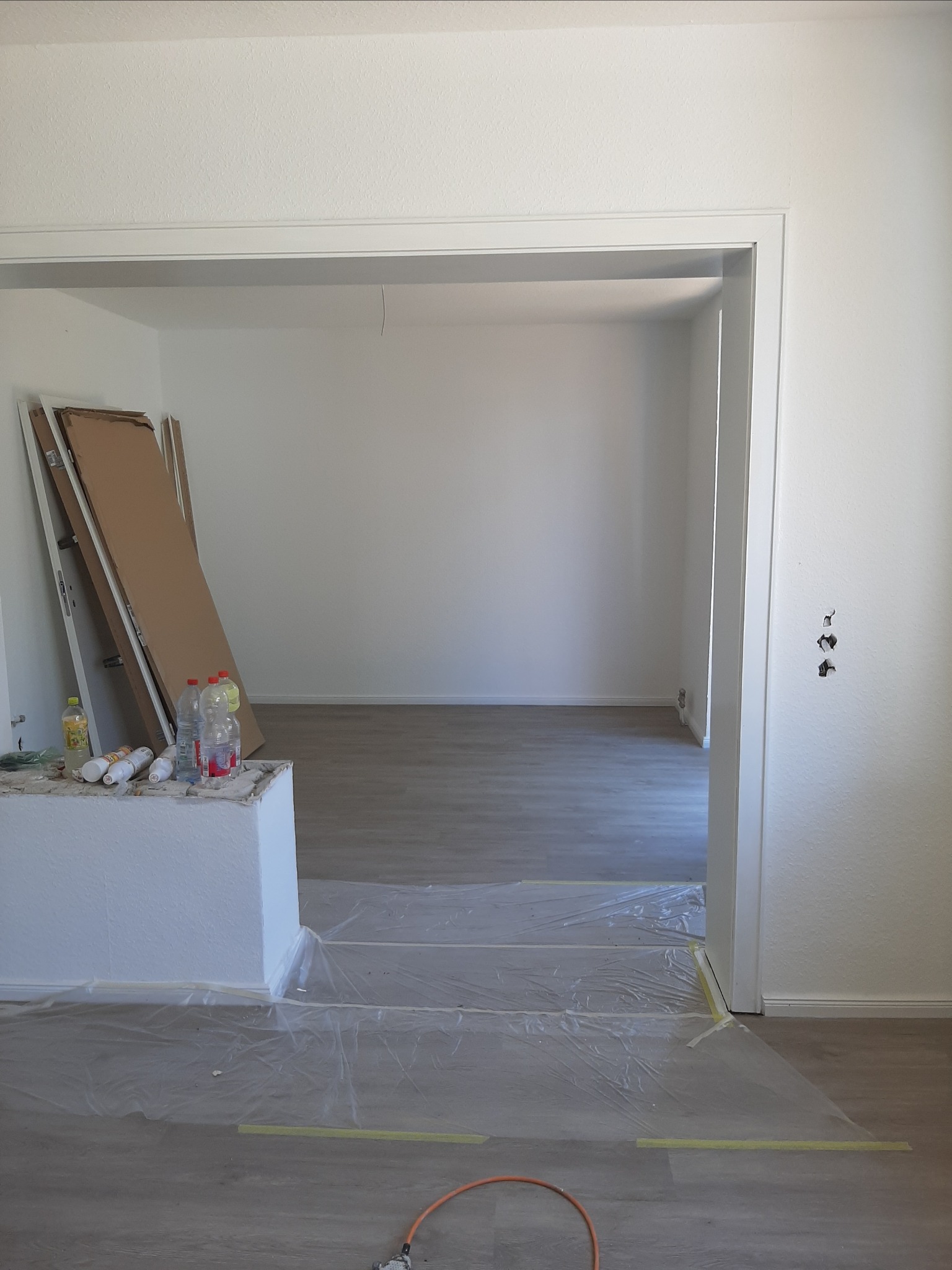 Przygotowanie do malowania: pokój z zabezpieczoną folią podłogą, widoczne farby, butelki z wodą, kartony przy ścianie, oraz puszki z farbą na murku.