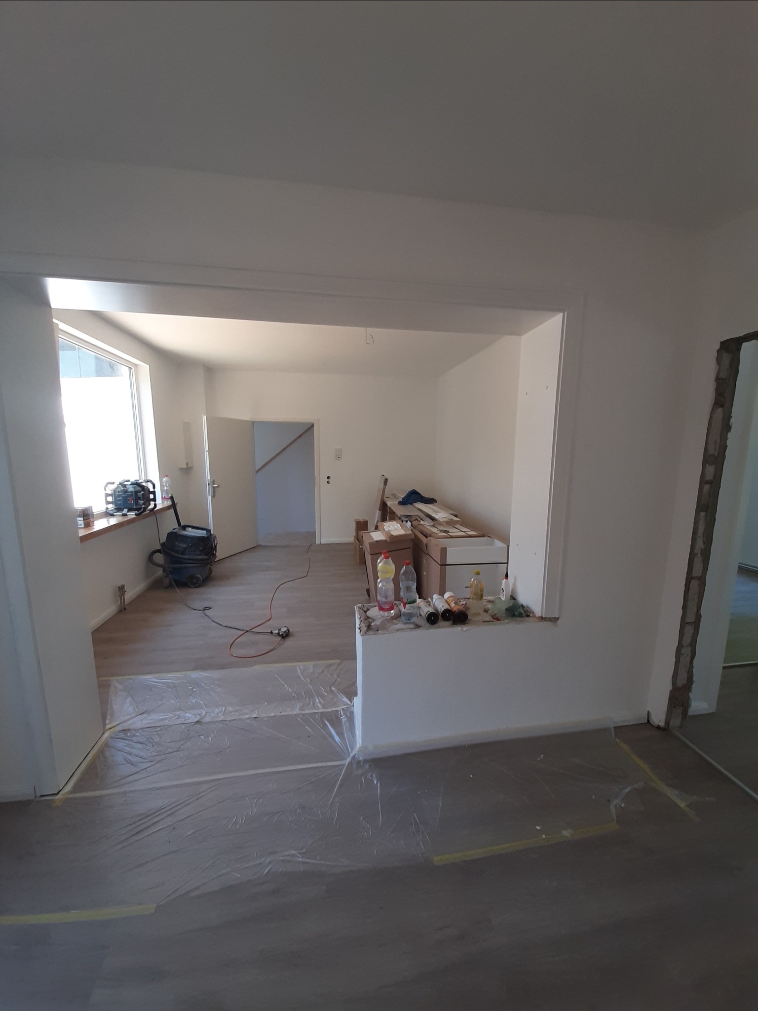 Przygotowanie do prac wykończeniowych: podłoga zabezpieczona folią malarską, świeżo pomalowane ściany na biało, widoczny odkurzacz przemysłowy i materiały budowlane w głębi pomieszczenia.