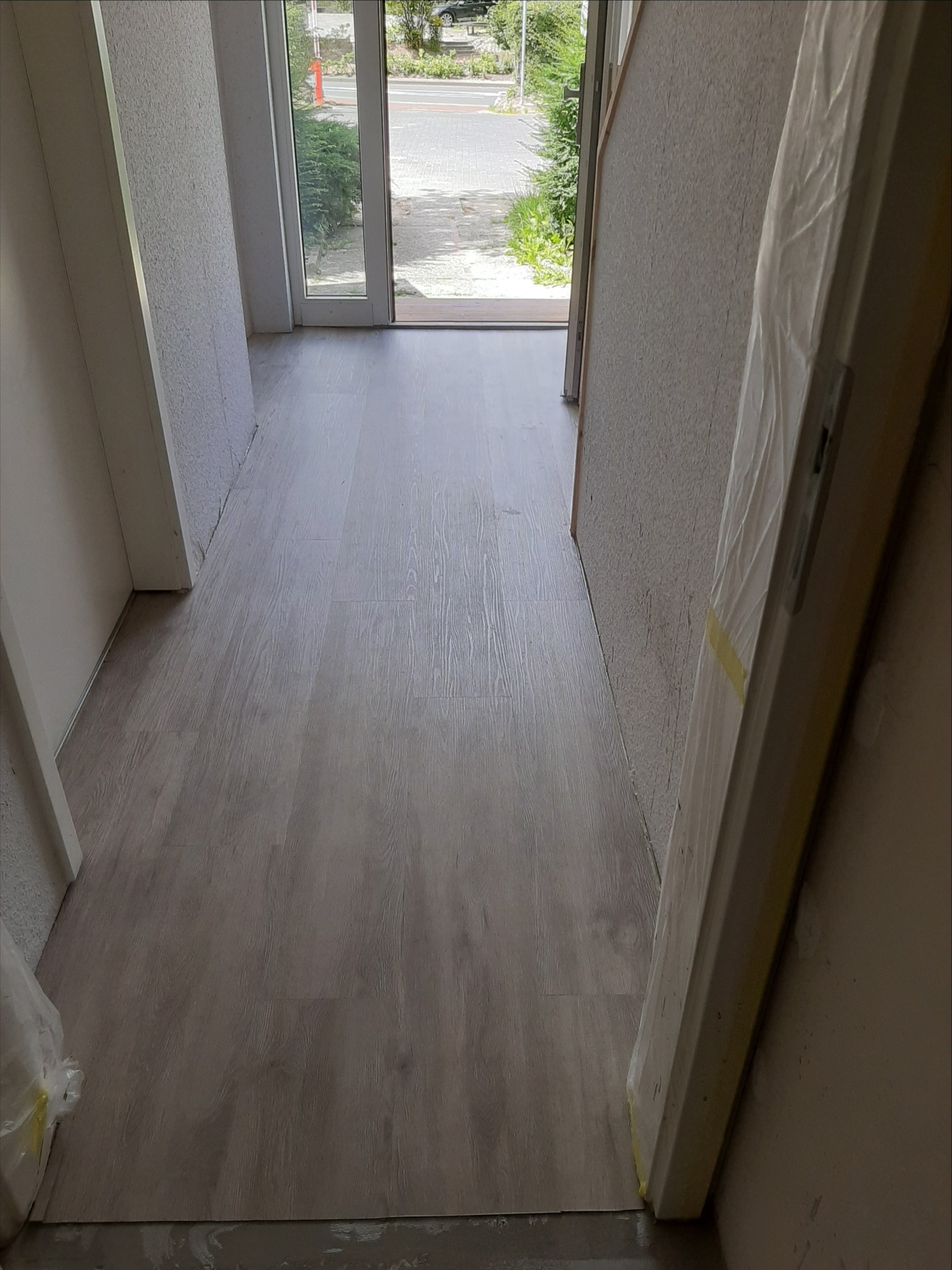 Wnętrze w trakcie prac wykończeniowych: świeżo położona podłoga z paneli w odcieniach szarości, surowe ściany i zabezpieczone folią drzwi wejściowe z widokiem na ulicę.