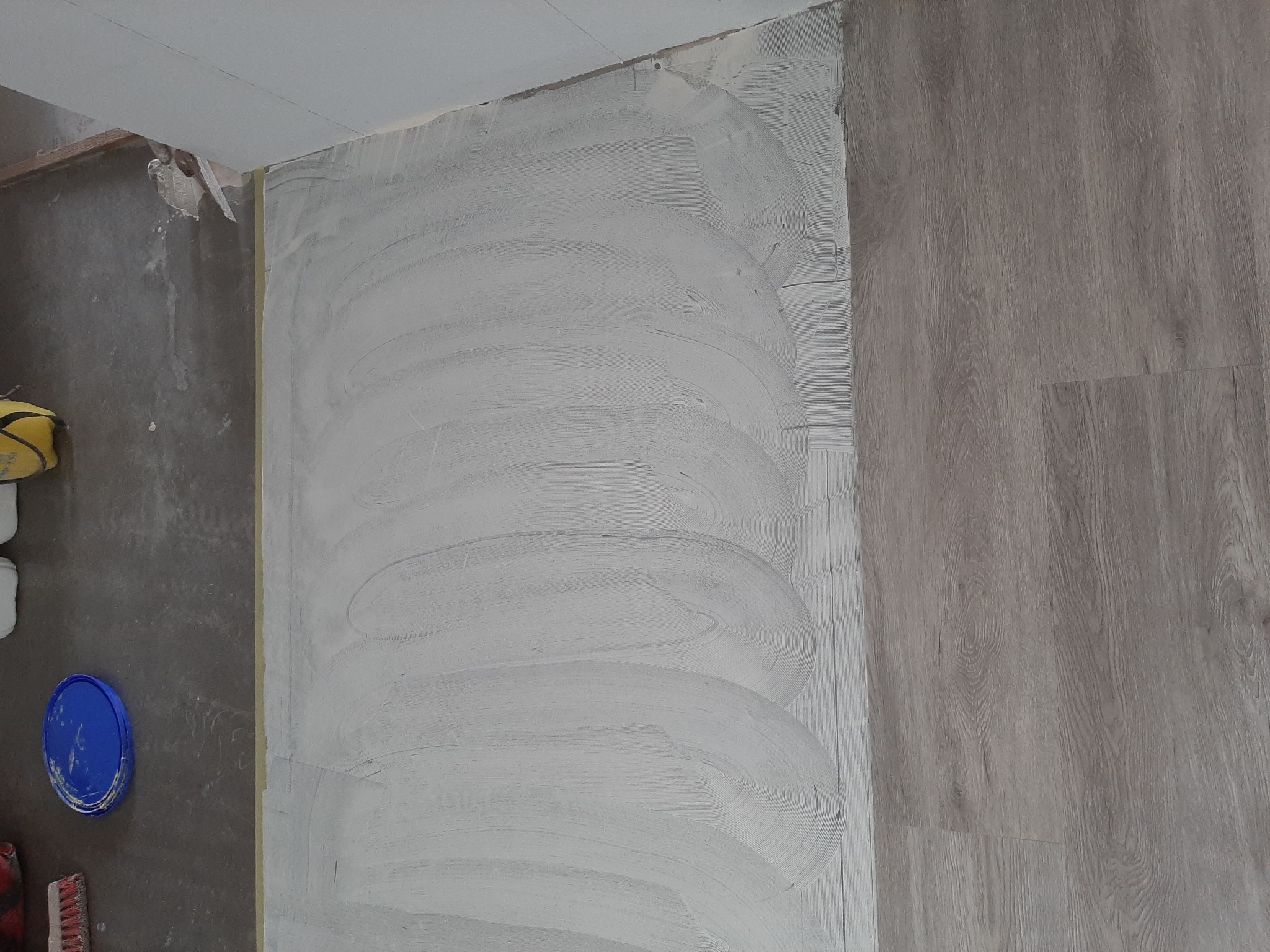 Świeżo nałożona warstwa gładzi na ścianie z widocznymi śladami po szpachli, fragment drewnopodobnej podłogi, wiadro z niebieską pokrywką i narzędzia malarskie w rogu.