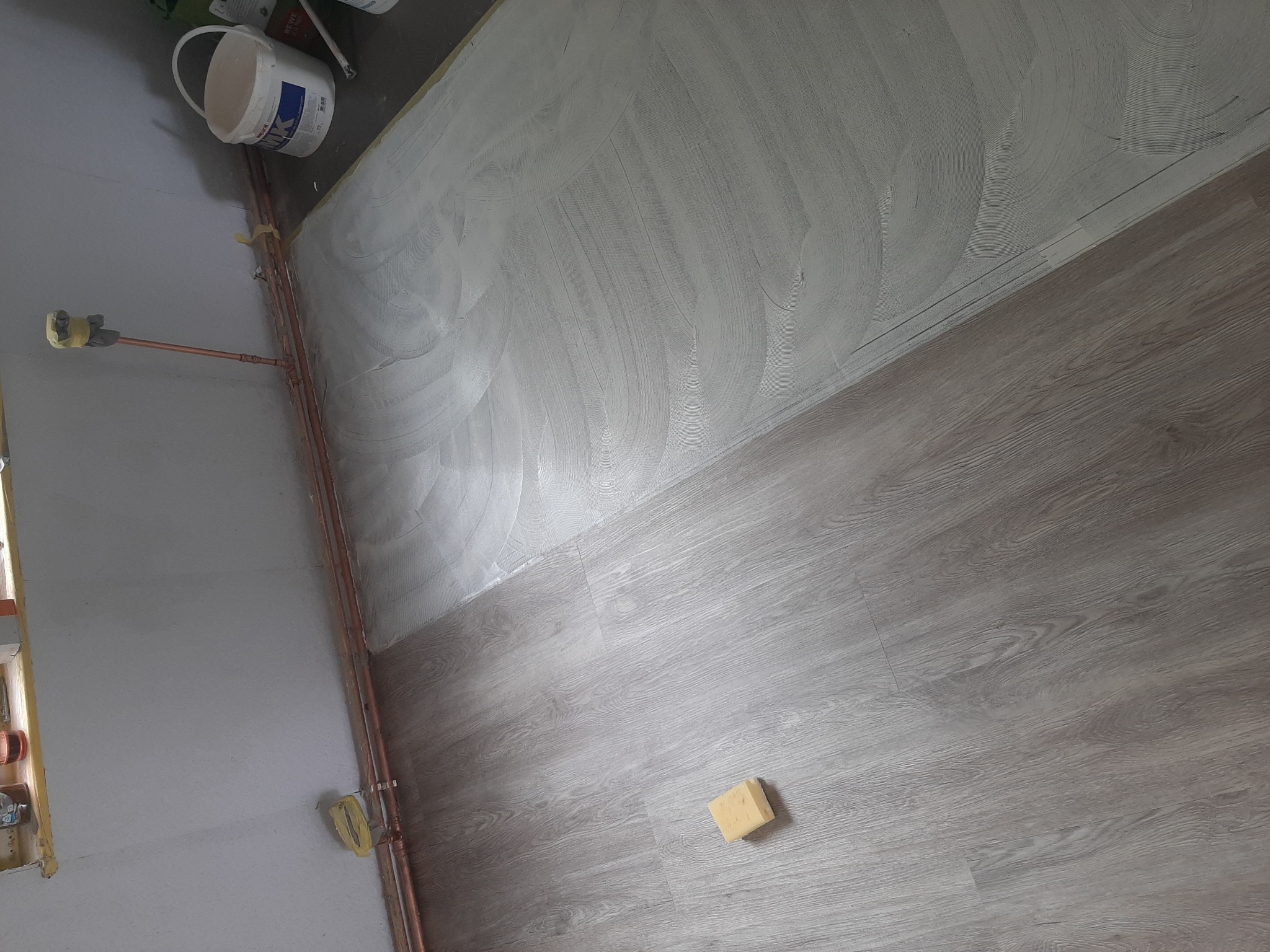 Świeżo nałożony, jasnoszary tynk maszynowy na ścianie, kontrastujący z panelami podłogowymi imitującymi drewno; widoczny fragment instalacji hydraulicznej i gąbka do czyszczenia.