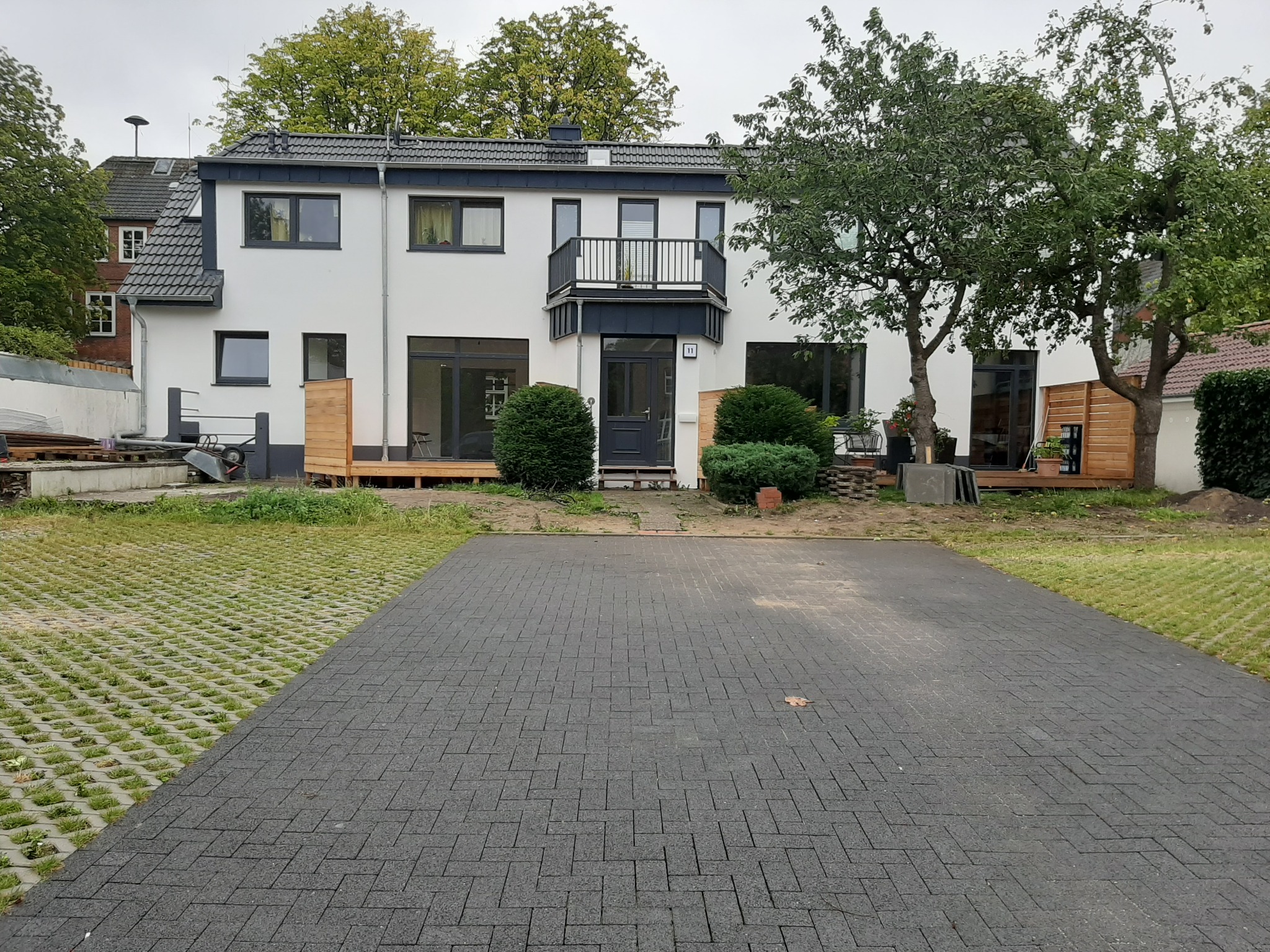 Biały dom z szarymi elementami wykończeniowymi, balkonem i numerem 11 nad wejściem, otoczony zielenią i częściowo wykończonym podjazdem z szarej kostki.