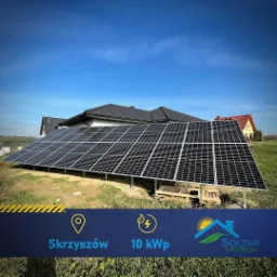 Instalacja fotowoltaiczna na gruncie przed domem jednorodzinnym, widoczne panele słoneczne na metalowej konstrukcji, w tle budynek mieszkalny z ciemną dachówką, błękitne niebo.