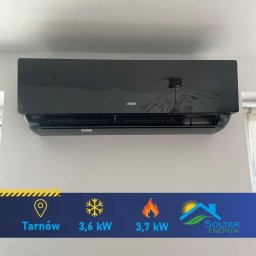 Soltar Energia Sp. z o.o. - Czarny klimatyzator AUX na białej ścianie, widoczne parametry techniczne i logo Soltar Energia. Odbicie lampy w obudowie. Nowoczesny design.