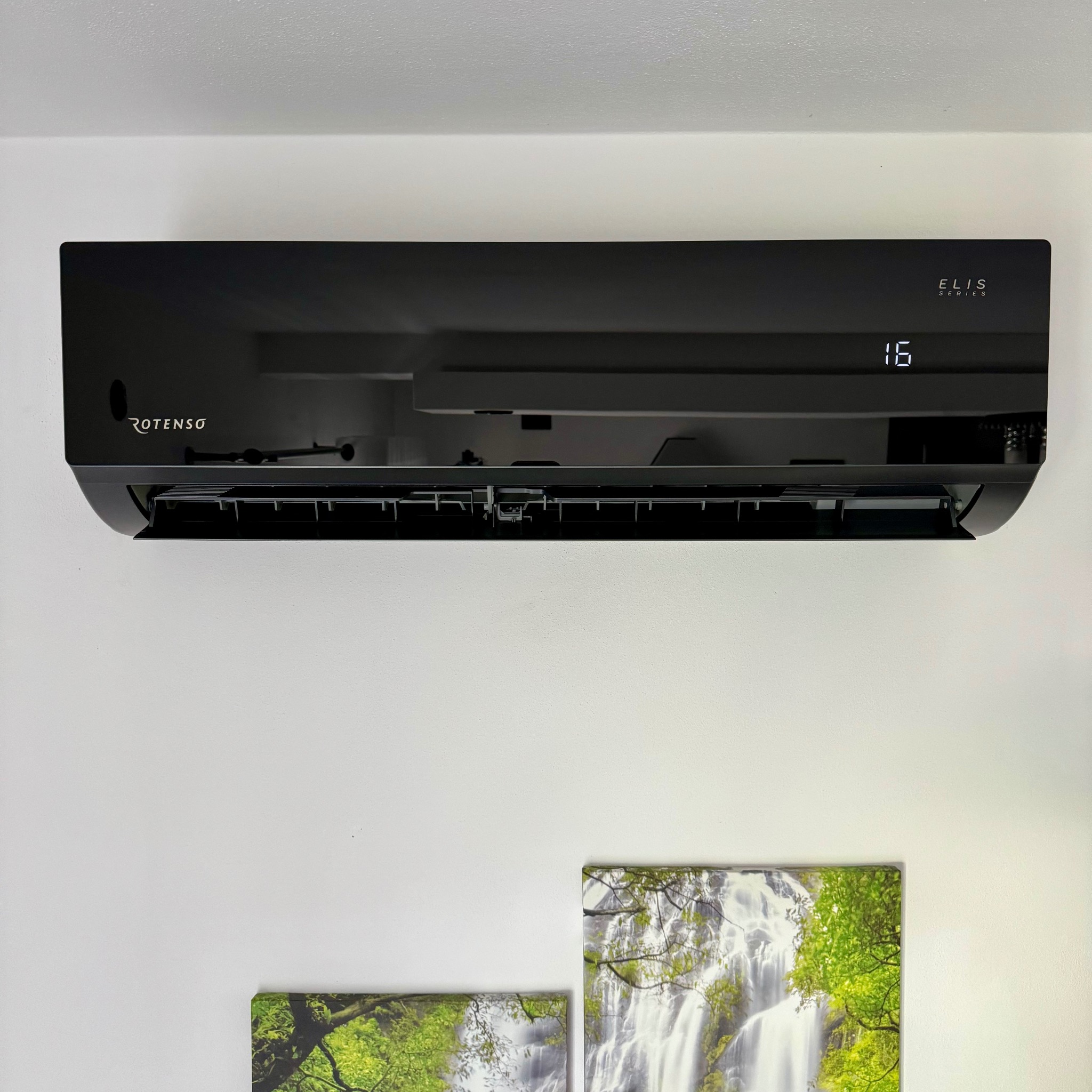 Czarny klimatyzator Rotenso Elis Series na białej ścianie, z wyświetlaczem temperatury 16 stopni i dwoma obrazami z motywem wodospadu poniżej.