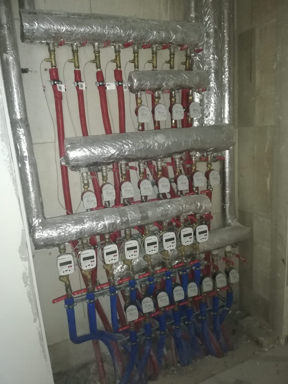 Rozdzielacz ogrzewania podłogowego z licznikami ciepła, zaizolowany folią aluminiową, zamontowany w Karnieszewicach. Widoczne czerwone i niebieskie rury.