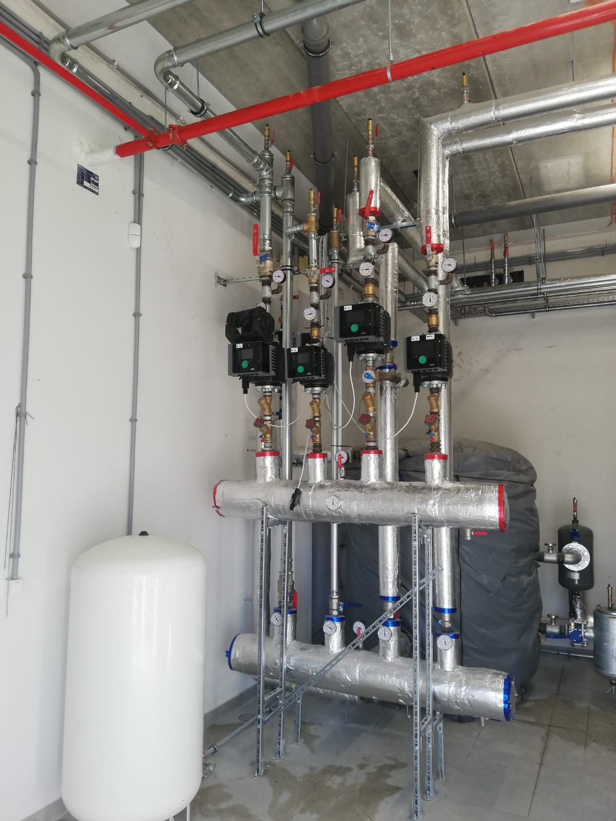Węzeł cieplny w kotłowni: biały zbiornik wyrównawczy, zaizolowane rury, zawory, manometry, pompy obiegowe, instalacja przeciwpożarowa z rurami w kolorze czerwonym.