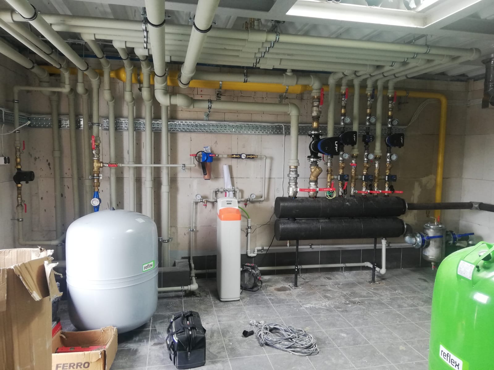 Złożona instalacja centralnego ogrzewania w kotłowni z licznymi rurami, zaworami, pompami obiegowymi i zbiornikami wyrównawczymi Reflex, widoczne również elementy uzdatniania wody.