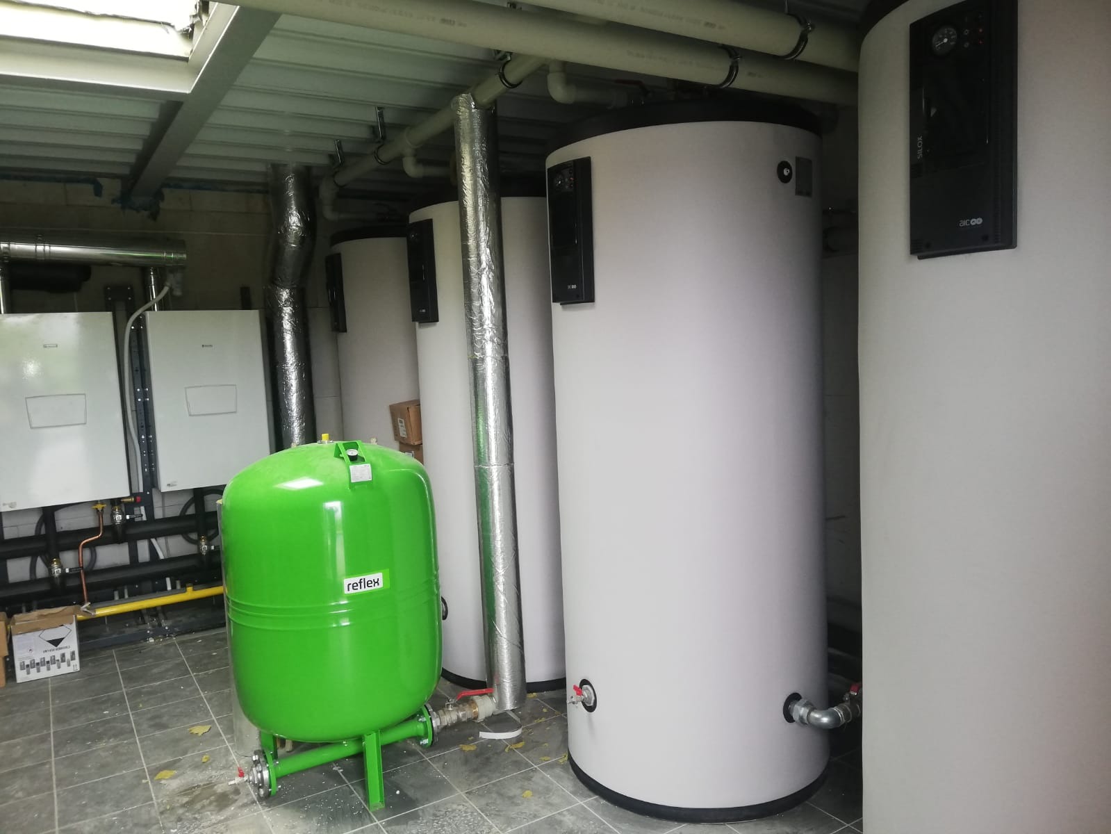Instalacja kotłowni z zielonym zbiornikiem przeponowym Reflex, białymi zasobnikami ciepłej wody i dwoma kotłami grzewczymi na ścianie.