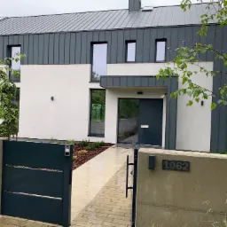 Nowoczesny dom jednorodzinny z grafitowym dachem z blachy na rąbek stojący i elewacją w kolorze złamanej bieli, widok z poziomu chodnika z kostki brukowej, brama wjazdowa z numerem posesji 1062.