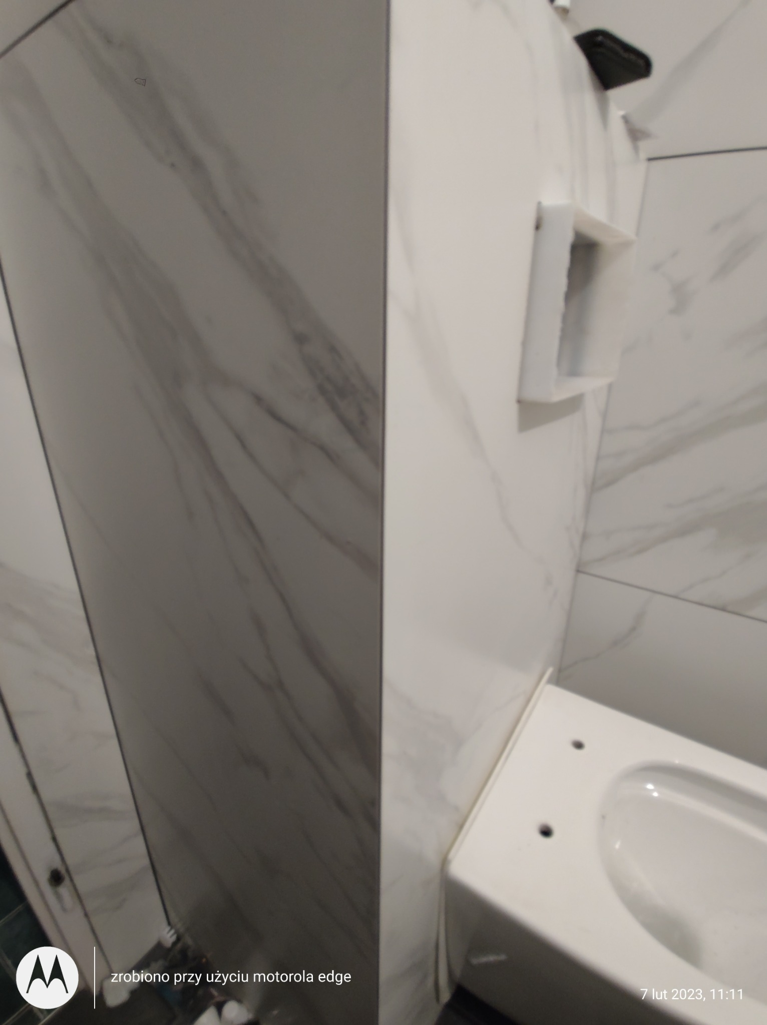 Fragment łazienki w trakcie wykańczania: białe płytki imitujące marmur, częściowo zamontowana toaleta, widoczna instalacja podtynkowego stelaża WC.