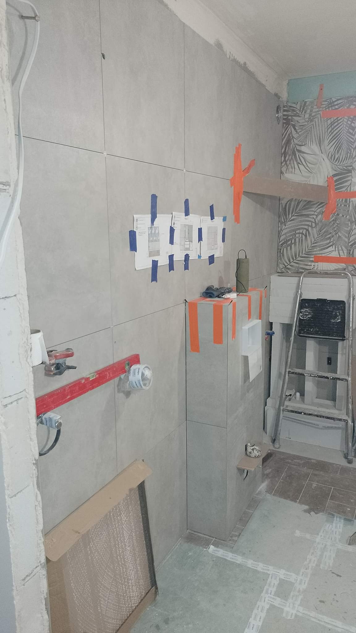 Łazienka w trakcie remontu: szare płytki na ścianach, poziomica laserowa, kartki z planami przyklejone taśmą, drabina aluminiowa, częściowo ułożone płytki podłogowe.
