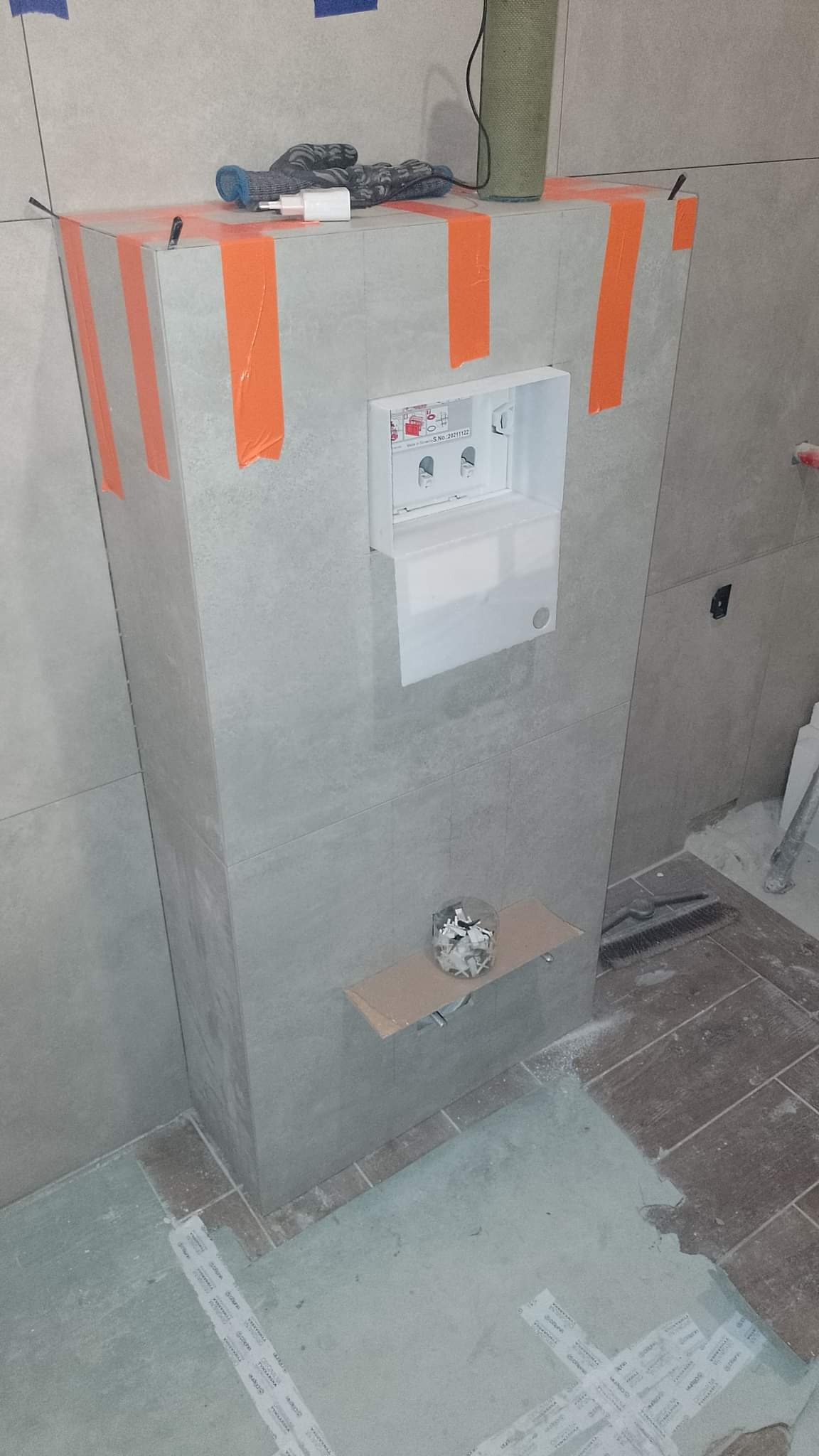 Szara zabudowa stelaża WC w trakcie prac wykończeniowych, zabezpieczona pomarańczową taśmą, z widocznym pojemnikiem na przyciski spłukujące i akcesoriami na półce, rękawice robocze i ładowarka...