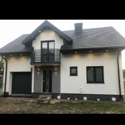 Dwupiętrowy dom z szarą elewacją, czarnym dachem i balkonem, oświetlony zewnętrznymi lampami, z widocznym garażem i dekoracyjnymi kulami w ogrodzie.