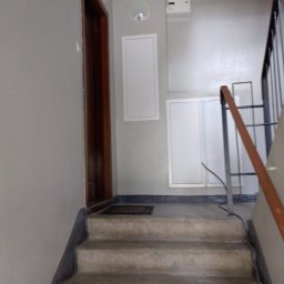 Drabex - Klatka schodowa w bloku po malowaniu ścian na szaro, widoczne schody z szorstką fakturą, drzwi wejściowe i poręcze schodów.