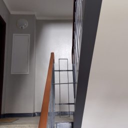 Drabex - Klatka schodowa w budynku mieszkalnym po odświeżeniu, widoczne pomalowane ściany w jasnych odcieniach szarości i beżu, schody z szorstką powierzchnią i metalowe barierki z drewnianym poręczą.