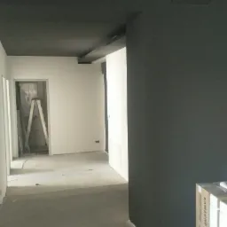 Wnętrze w trakcie wykańczania, z pomalowanym na ciemny kolor sufitem i fragmentem ściany, w tle drabina oparta o ścianę, na podłodze widoczne ślady po pracach remontowych, kartonowe opakowanie przy...