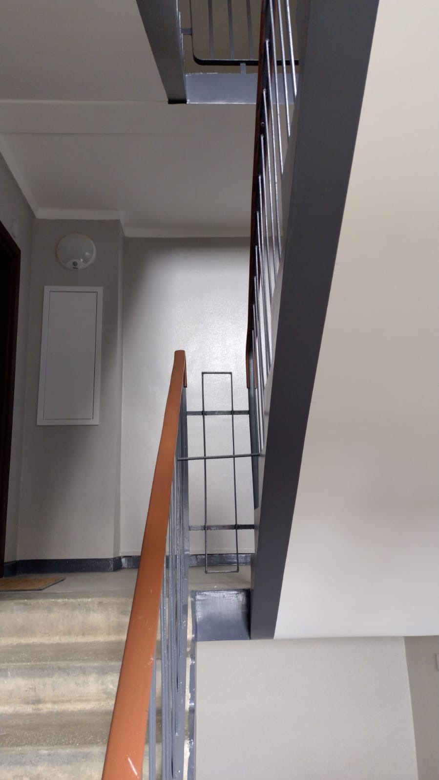 Klatka schodowa w budynku mieszkalnym po odświeżeniu, widoczne pomalowane ściany w jasnych odcieniach szarości i beżu, schody z szorstką powierzchnią i metalowe barierki z drewnianym poręczą.