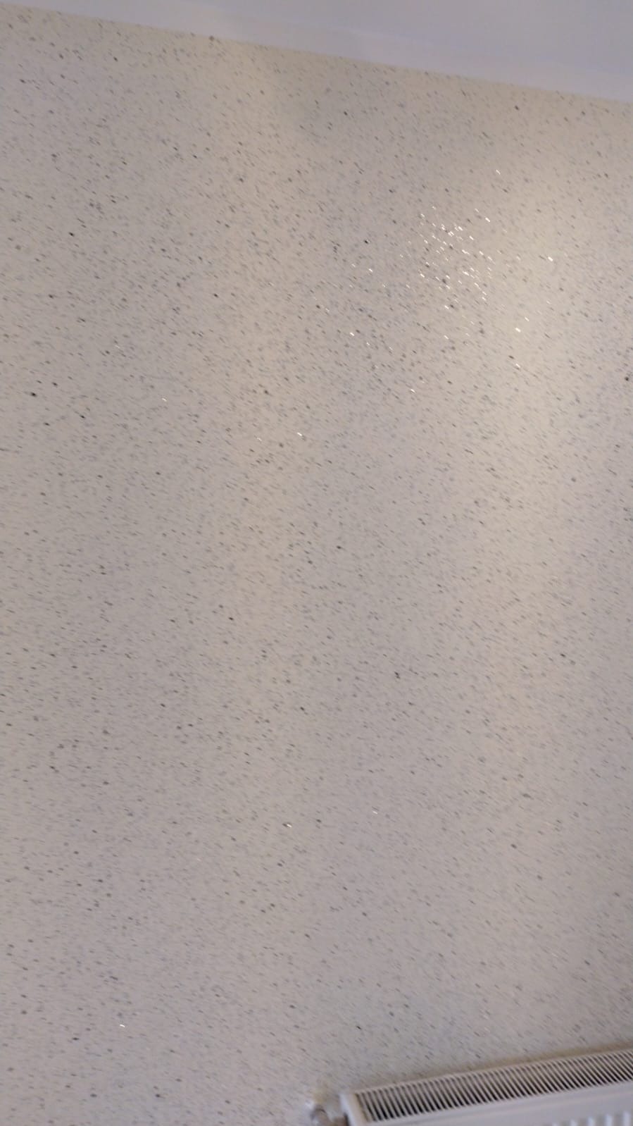 Jasnoszara ściana wykończona szpachlą z drobnymi, ciemnymi punktami, widoczne odbicia światła, u dołu fragment grzejnika.
