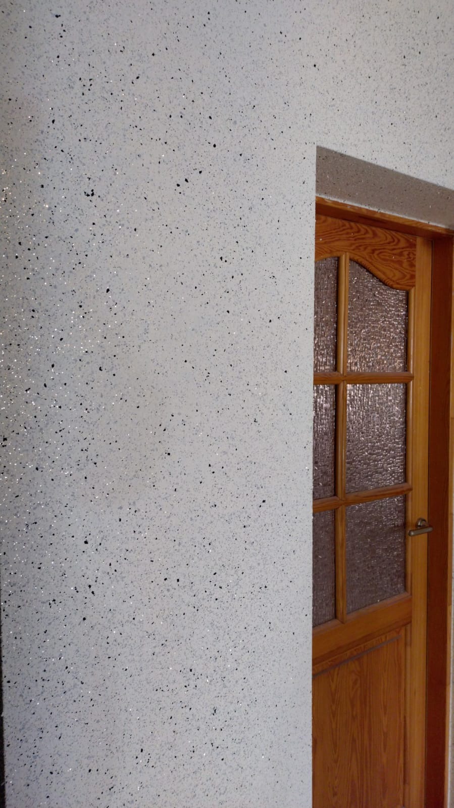 Ściana wewnętrzna pokryta białą, dekoracyjną strukturą z czarnymi i srebrnymi drobinkami, sąsiadująca z drewnianymi drzwiami z szybą o fakturze szkła ornamentowego.