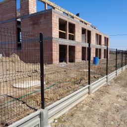 Tomstal - Czarny panelowy system ogrodzeniowy z podmurówką betonową na tle budowy domu z czerwonej cegły, widoczny błękitny pojemnik i pracownik na dachu.