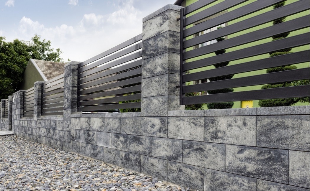 Nowoczesne ogrodzenie z poziomych, czarnych profili metalowych na niskiej podmurówce z szarej kostki betonowej imitującej kamień, widok z poziomu gruntu na tle zieleni.