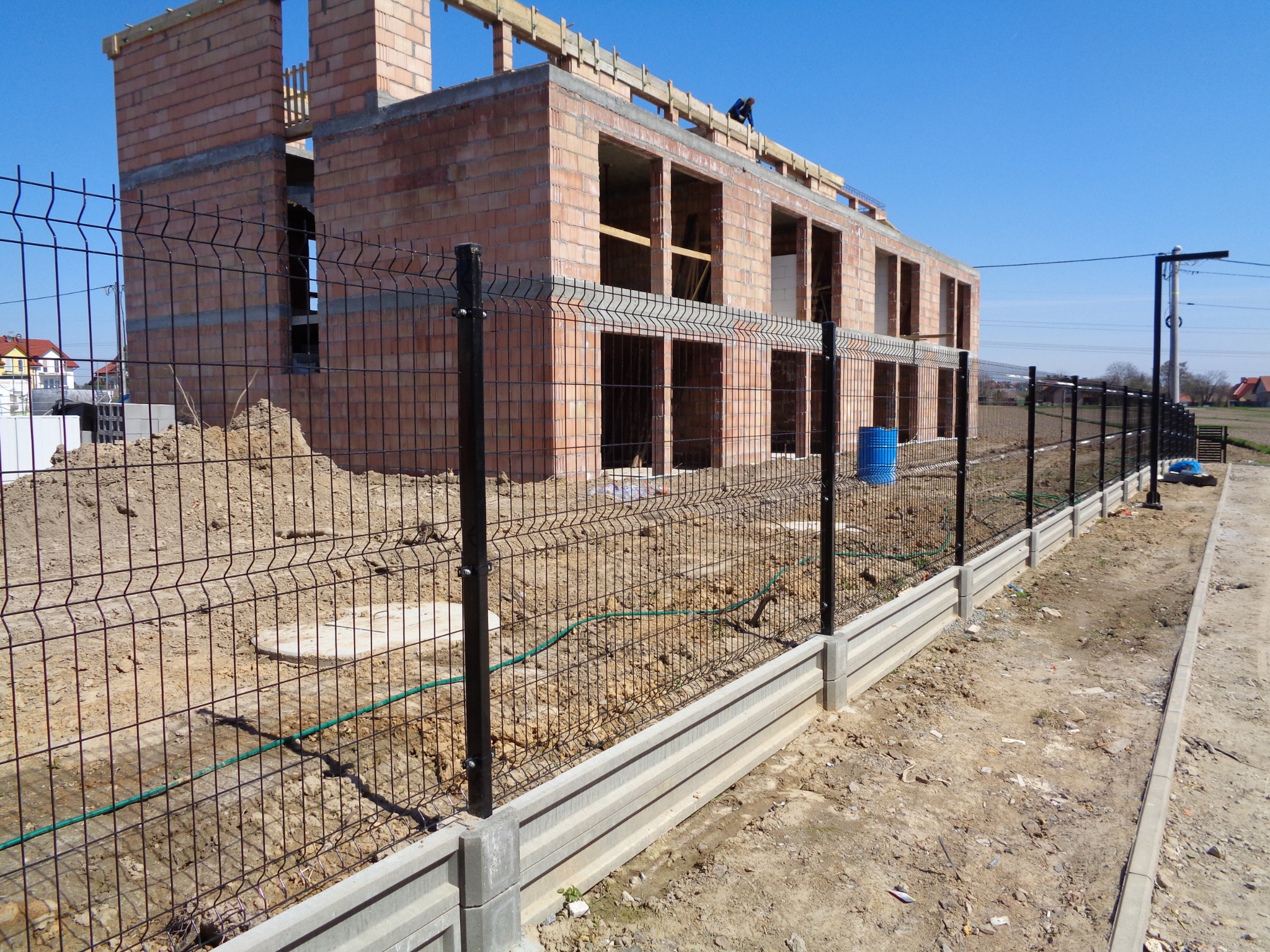 Czarny panelowy system ogrodzeniowy z podmurówką betonową na tle budowy domu z czerwonej cegły, widoczny błękitny pojemnik i pracownik na dachu.