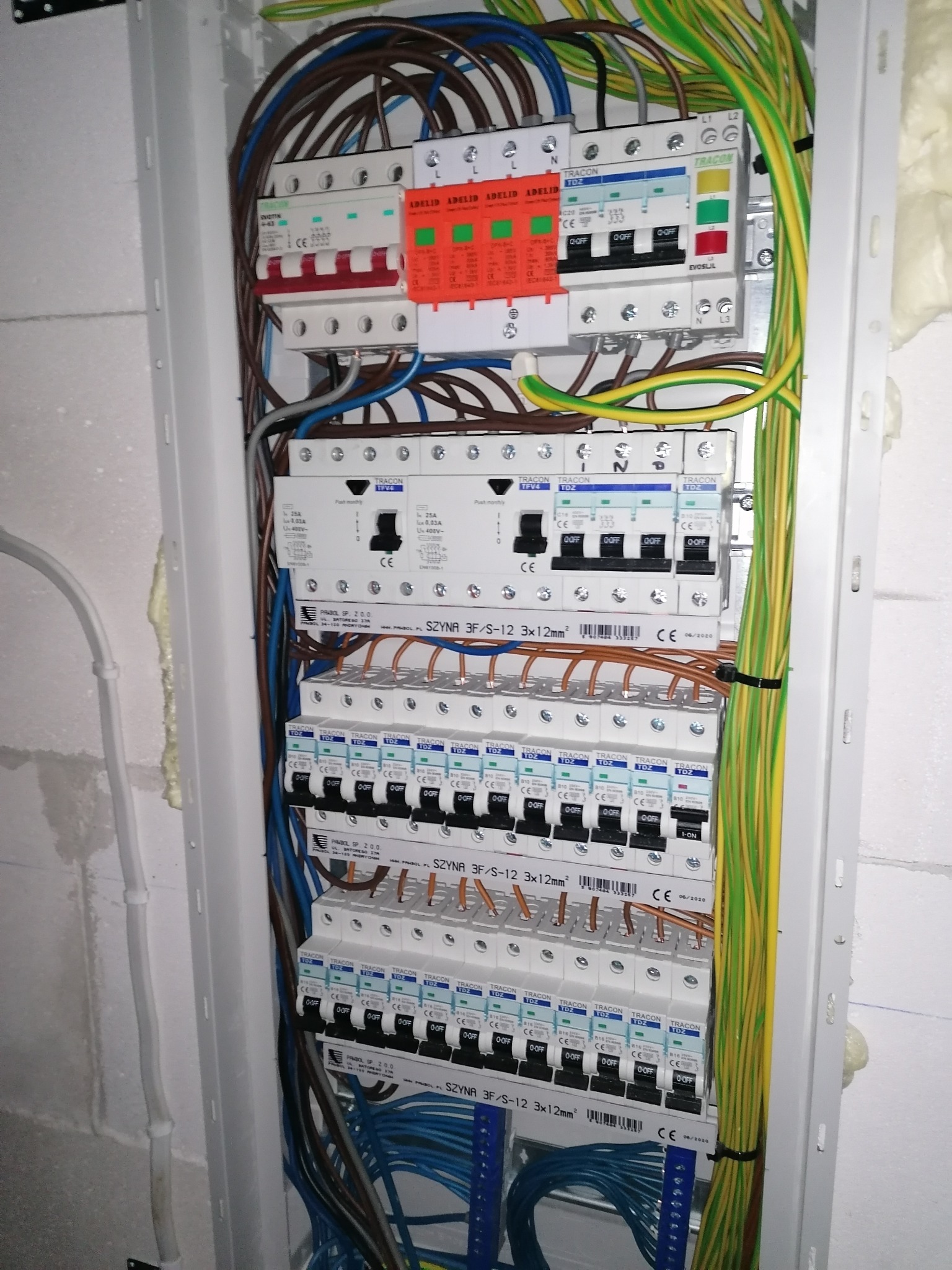 Rozdzielnica elektryczna z zainstalowanymi bezpiecznikami, przewodami i listwami zaciskowymi wewnątrz obudowy, widoczne różne modele i marki zabezpieczeń.