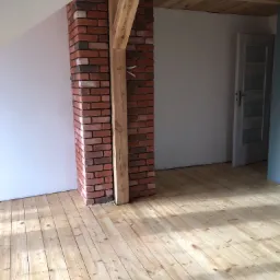 Wnętrze poddasza z drewnianą podłogą, ceglanym filarem obudowanym drewnianą konstrukcją oraz drewnianym sufitem, w tle białe ściany i drzwi.