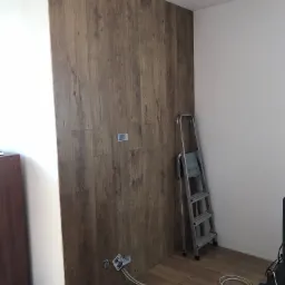 Ściana wyłożona panelami drewnopodobnymi w trakcie montażu, widoczne puszki elektryczne bez osprzętu i aluminiowa drabina oparta o ścianę.