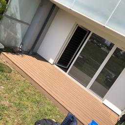 M&H Usługi remontowo-budowlane - Nowo wybudowany taras z desek kompozytowych, przylegający do nowoczesnego domu z dużymi oknami przesuwnymi, z widocznymi śladami budowy w postaci worka z odpadami i plecaka obok.