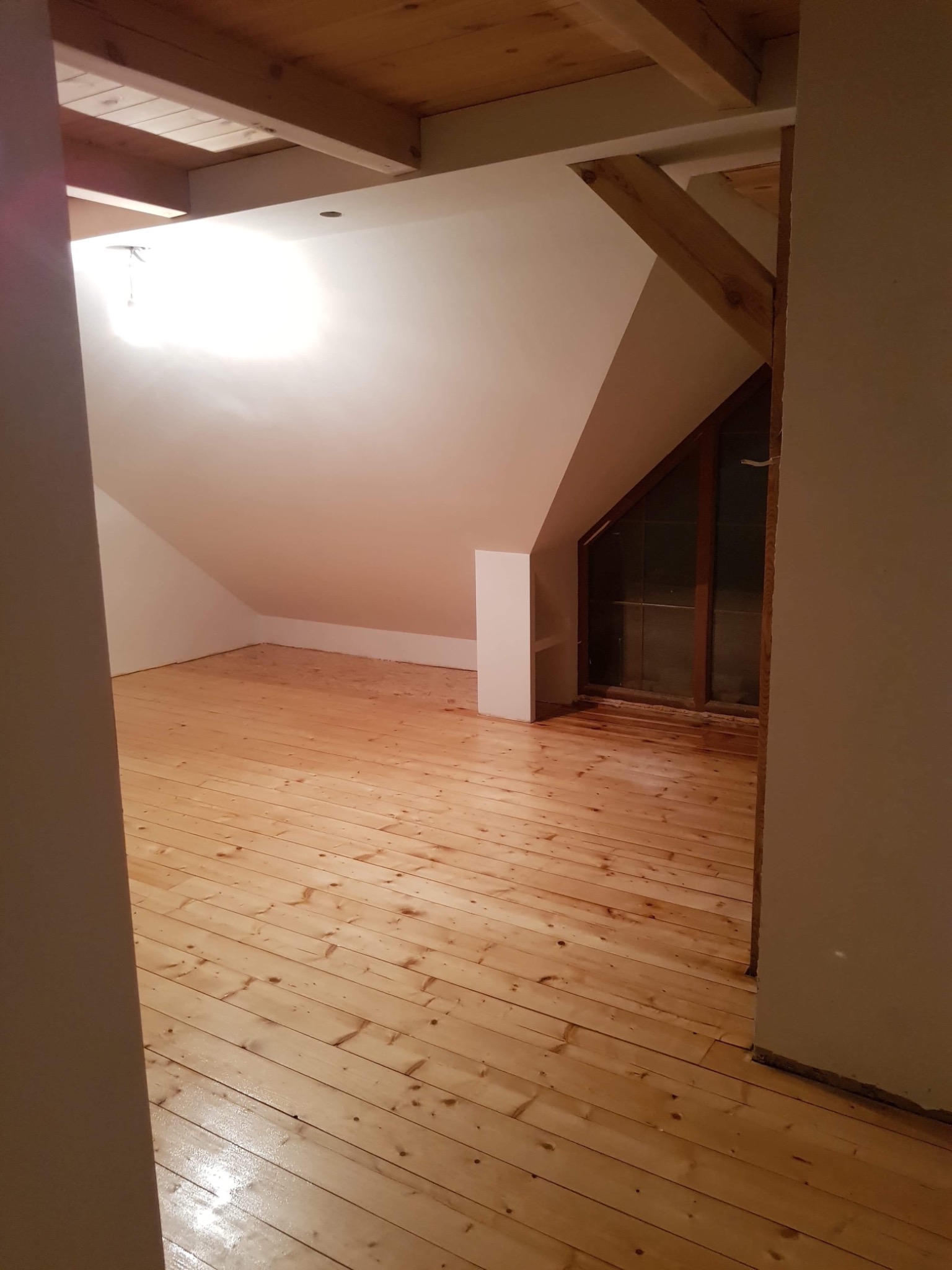 Poddasze z drewnianą podłogą po lakierowaniu, widoczne skosy ścian, drewniane belki stropowe i okno w szczycie, częściowo wykończone.