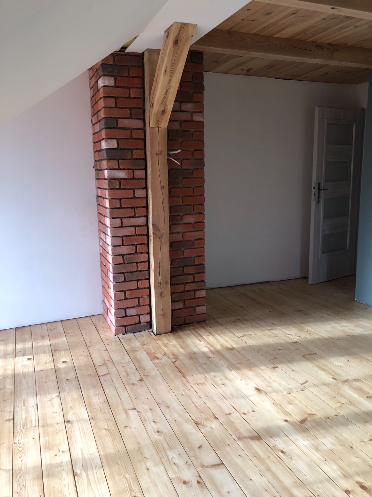 Wnętrze poddasza z drewnianą podłogą, ceglanym filarem obudowanym drewnianą konstrukcją oraz drewnianym sufitem, w tle białe ściany i drzwi.