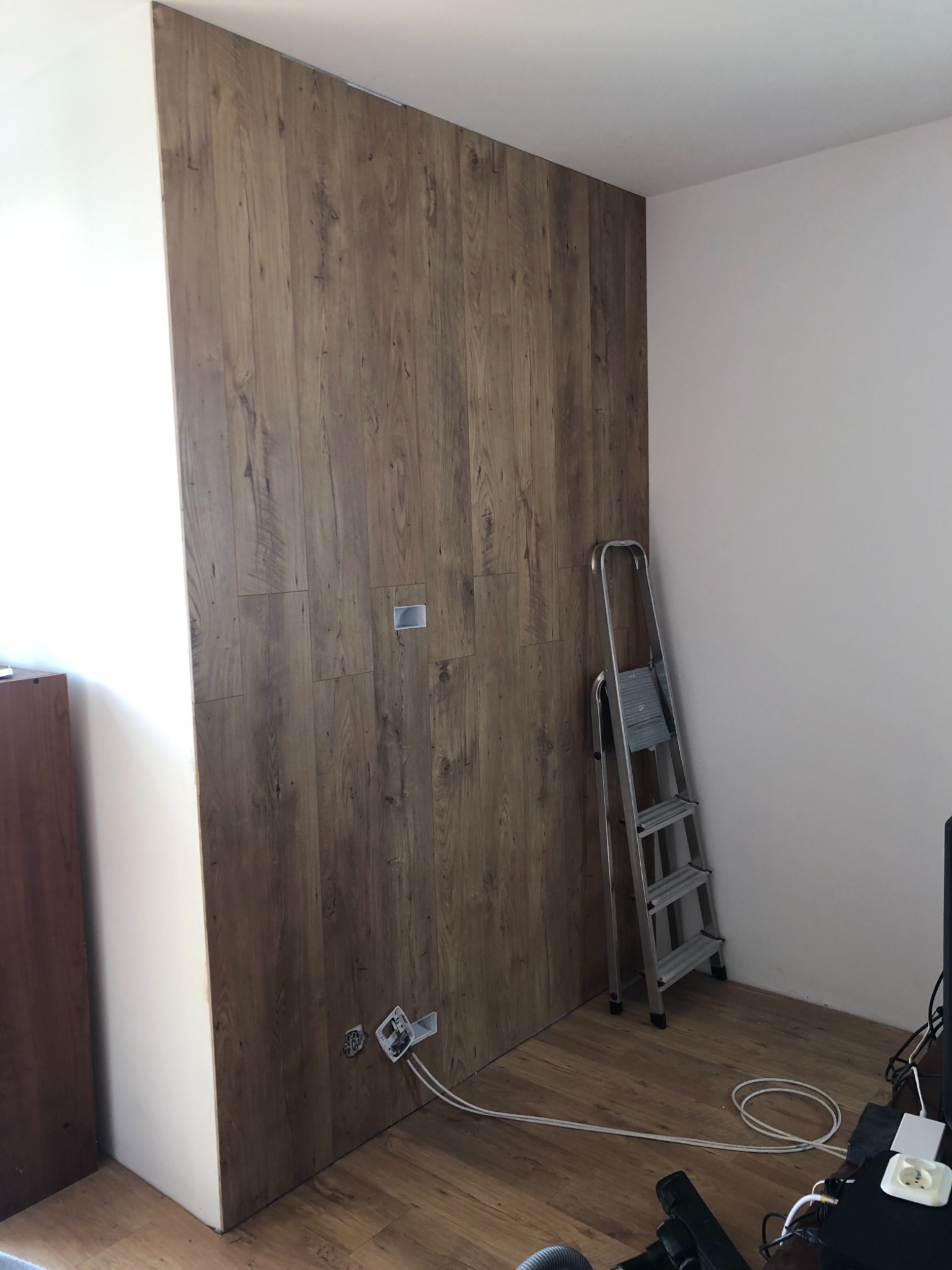Ściana wyłożona panelami drewnopodobnymi w trakcie montażu, widoczne puszki elektryczne bez osprzętu i aluminiowa drabina oparta o ścianę.